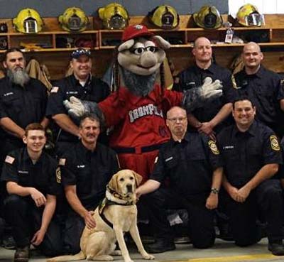 Yellow lab sits in front of a group of fireman and a mascot as they pose for a picture.