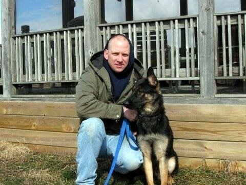 Man in winter coat and blue jeans kneels next to German Shephered.